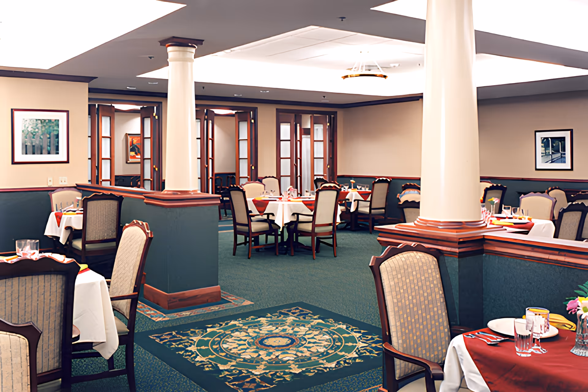 A formal dining room with multiple tables set with white tablecloths, red placemats, glassware, and silverware. The room features green carpet with a decorative pattern, beige walls with framed artwork, wooden trim, and large white columns. Chairs with upholstered backs and seats surround the tables.