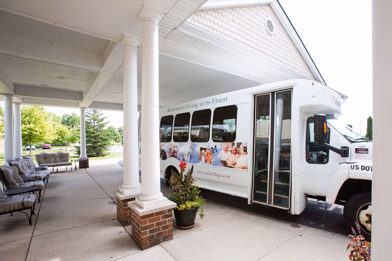 A white shuttle bus parked under a covered entrance with white columns and brick bases. The bus has images of elderly people and the text 'Retirement Living at its Finest' and 'www.seniorvillages.com' on its side. There are cushioned metal chairs and a bench along the left side under the covered area, with greenery and trees visible in the background.