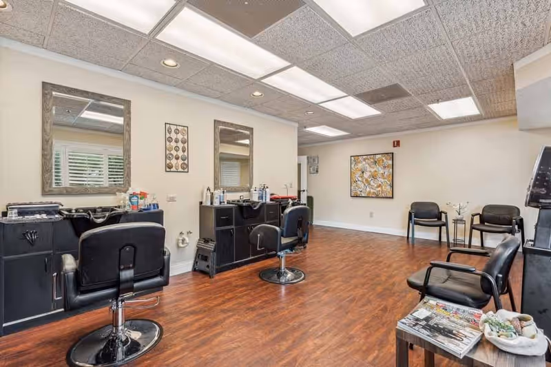 Interior view of a salon area with two black salon chairs in front of mirrors and counters with hair care products. The room has wood flooring, white walls, and ceiling lights. There are additional black chairs and a small table with magazines and decorative items in the corner.