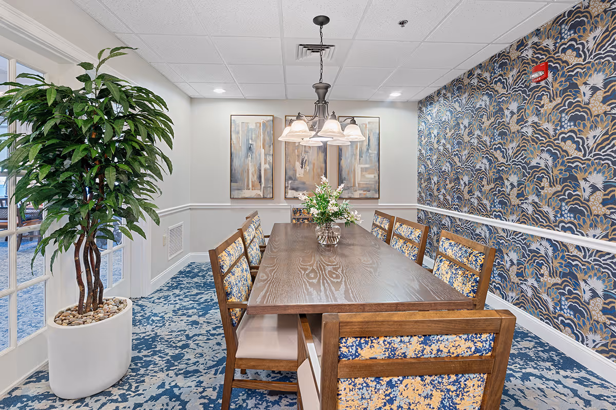 A dining room with a long wooden table surrounded by eight chairs with patterned upholstery. A vase with flowers is placed in the center of the table. The room features a blue and beige patterned carpet, a large potted plant near glass-paneled doors, abstract paintings on the wall, and a decorative wallpaper with a blue and gold design. A chandelier with multiple lights hangs above the table.