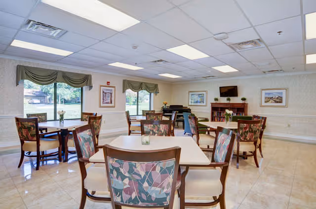 Bright communal dining/activity room with multiple tables and chairs, large windows, and a TV/bookcase.