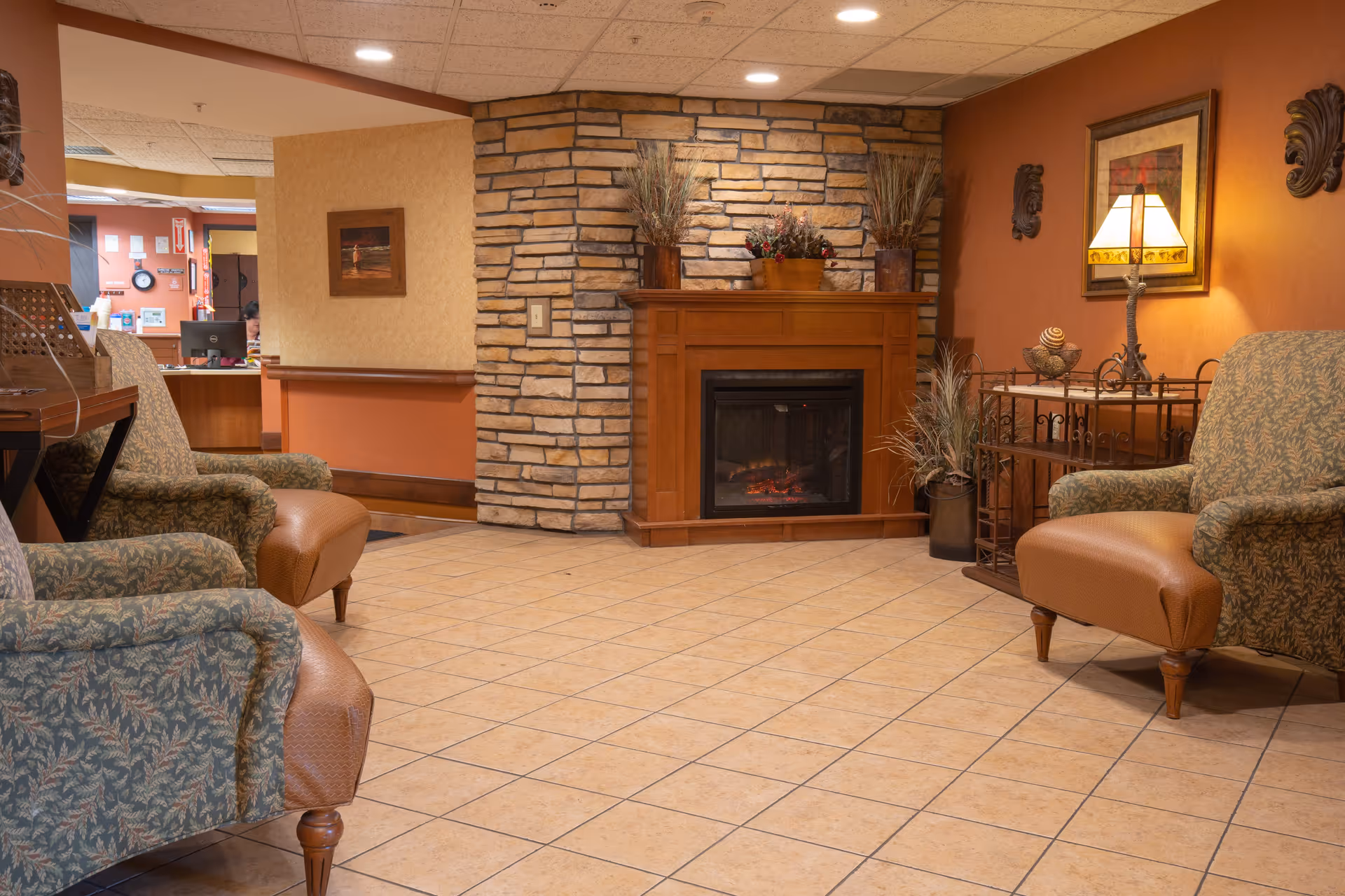 A cozy sitting area in a nursing and rehab facility featuring two upholstered armchairs with patterned fabric and leather footrests, a stone fireplace with a wooden mantle decorated with plants, a side table with a lamp and decorative items, warm-toned walls, and tiled flooring. In the background, a reception desk area is partially visible.