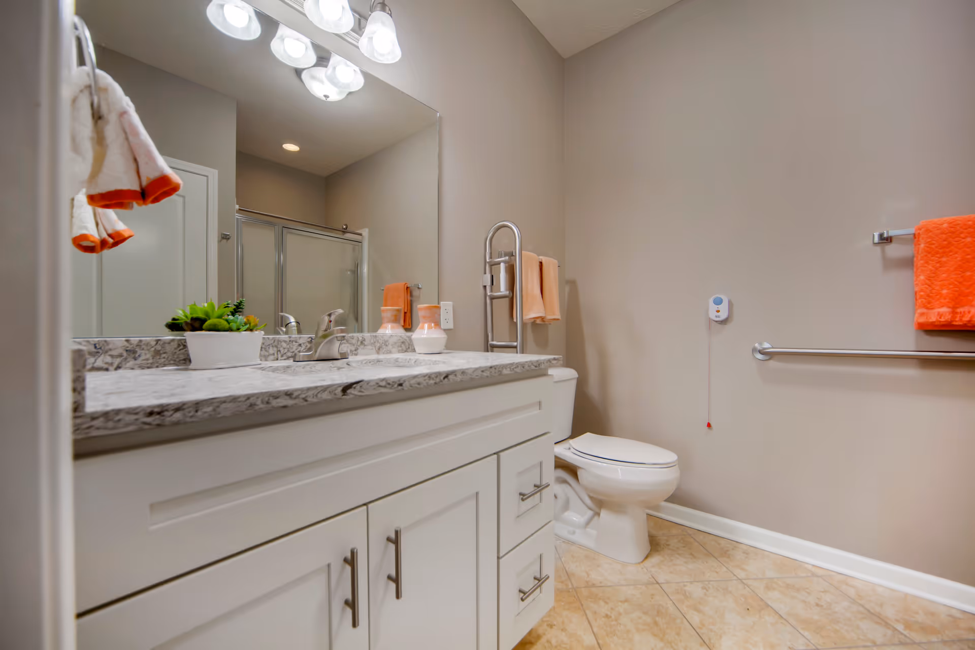 A clean and modern bathroom with beige walls and tiled floor. The bathroom features a white vanity with a marble countertop, a sink, and a large mirror above it. There are decorative items on the countertop including a small potted plant and two vases. A toilet with a safety grab bar is visible next to the vanity. Orange and peach-colored towels hang on towel racks, and a pull cord emergency alert device is mounted on the wall near the toilet. The shower area has a glass door.