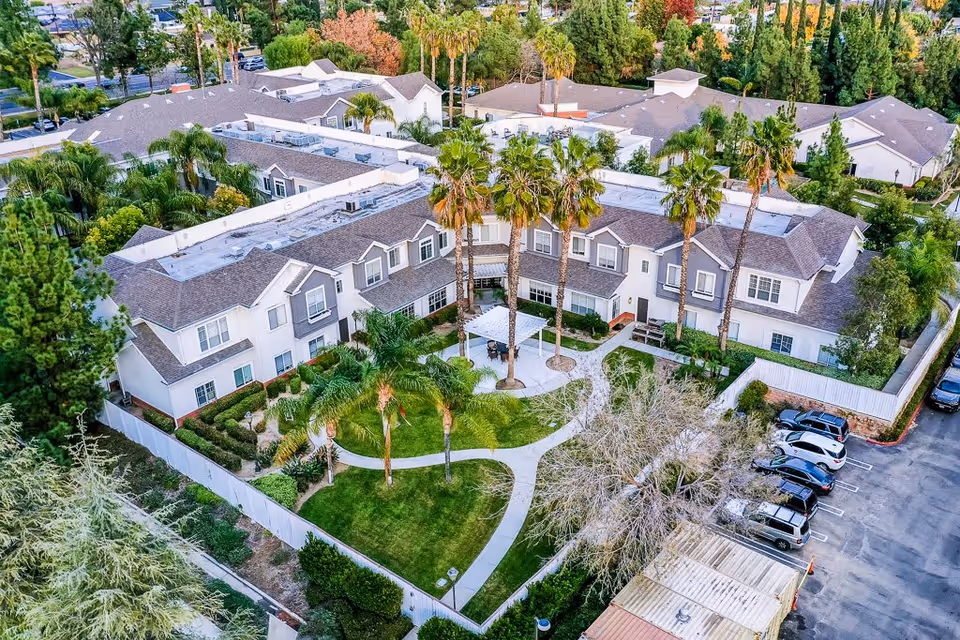 Aerial view of a senior living facility named Gardens At Northridge showing a two-story building surrounding a landscaped garden area with palm trees, walking paths, and a shaded seating area. There is a parking lot adjacent to the building with several parked cars.