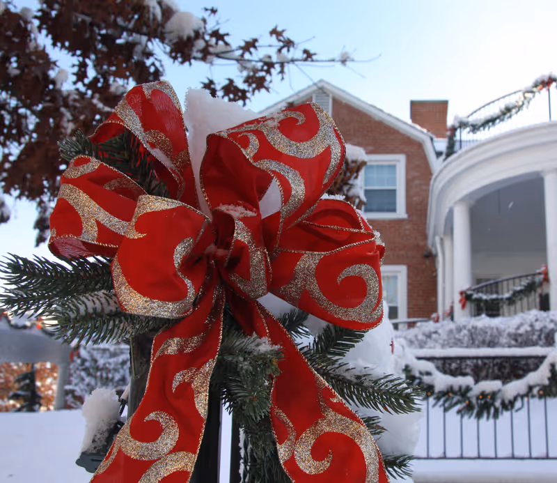 Close-up of a festive red and gold ribbon tied around a green garland with snow on it, in front of a snow-covered building with white columns and brick walls decorated with garlands.