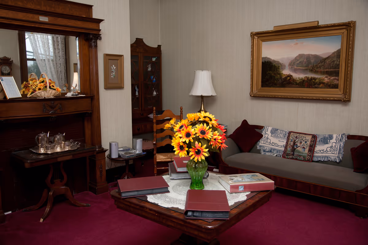 A cozy living room with a vintage wooden fireplace mantel, a green vase with bright yellow sunflowers on a wooden coffee table covered with a lace doily, several photo albums, a wooden chair, a gray couch with decorative pillows and a throw, a floor lamp, a glass-front cabinet, and a framed landscape painting on the wall.