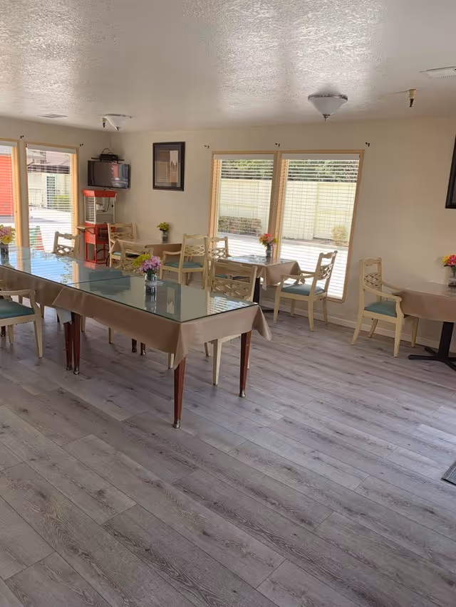 Bright dining area with several tables and chairs, a glass-topped central table, floral centerpieces, and large windows.