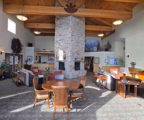 A spacious living room area with a high wooden beam ceiling and a large stone fireplace in the center. The room is furnished with multiple seating arrangements including sofas, armchairs, and a round wooden table with four wicker chairs. The space is well-lit with natural light coming from windows and ceiling lights. There are decorative plants and artwork on the walls, creating a warm and inviting atmosphere.