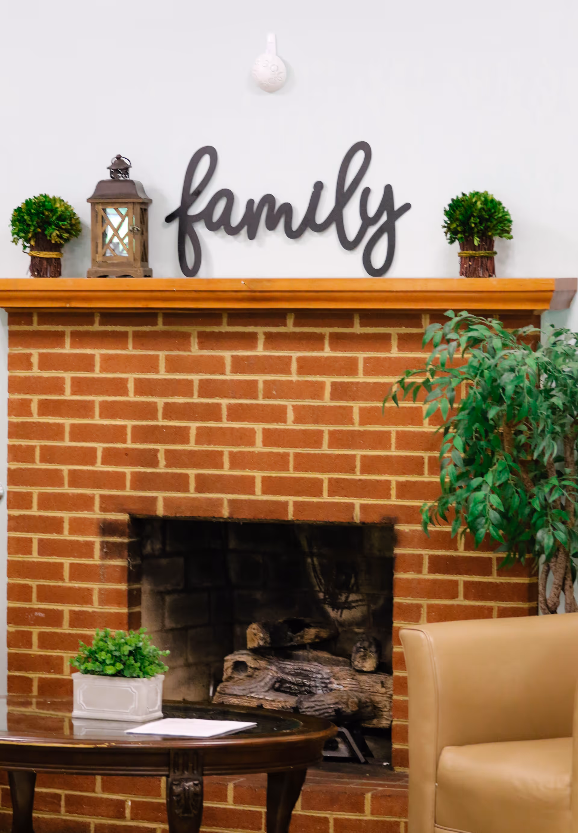 A cozy indoor scene featuring a brick fireplace with logs inside. Above the fireplace mantel is a decorative sign that says 'family' flanked by two small potted plants and a lantern. To the right of the fireplace is a green leafy plant and a beige armchair. In front of the fireplace is a wooden coffee table with a small potted plant and some papers on it.