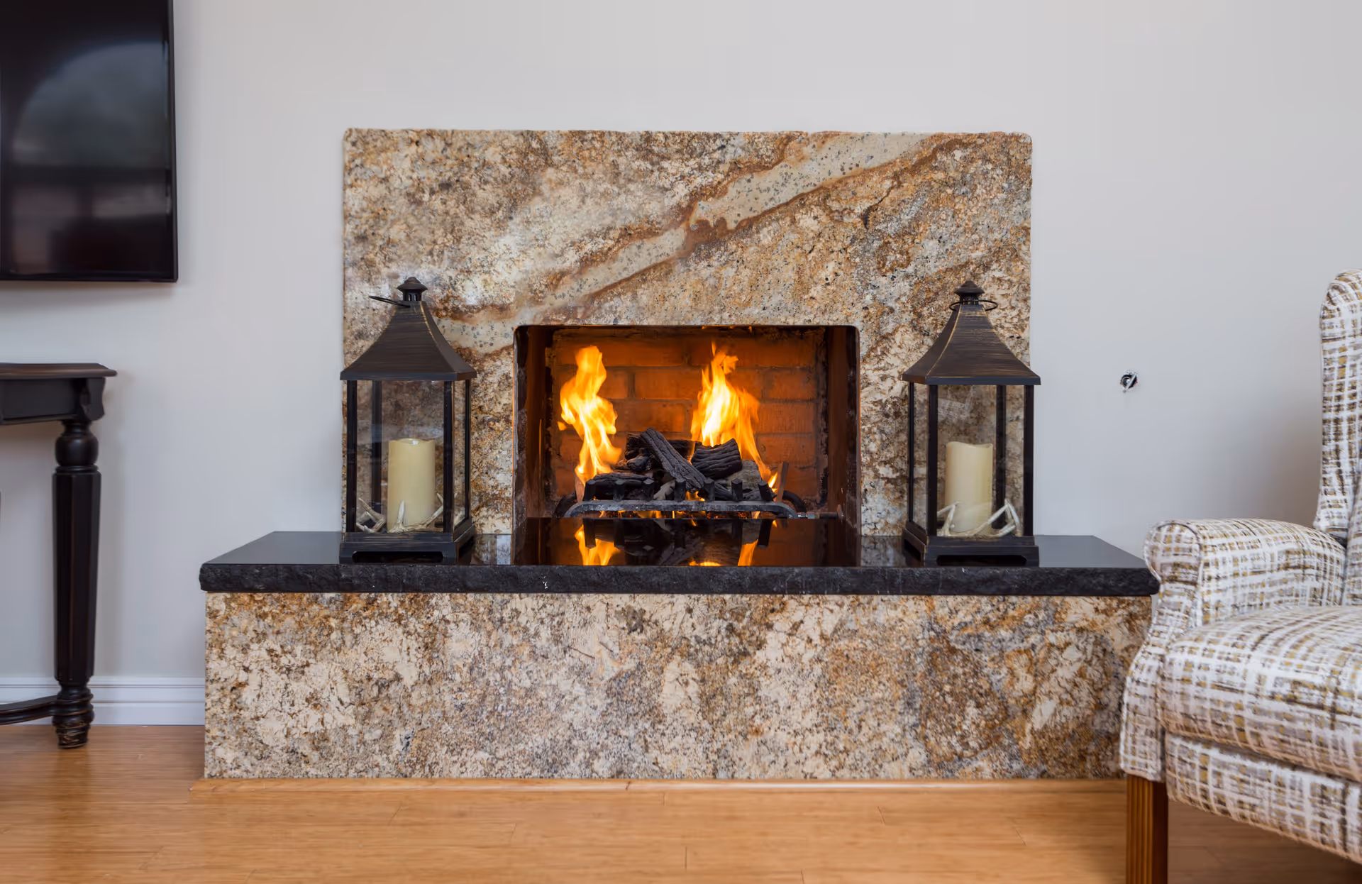 A cozy indoor fireplace with a granite surround and black mantel. Two decorative lanterns with candles are placed symmetrically on either side of the fireplace. To the right, part of a patterned upholstered armchair is visible, and to the left, a small dark wooden table and a mounted flat-screen TV are partially seen. The floor is wooden, and the walls are painted white.