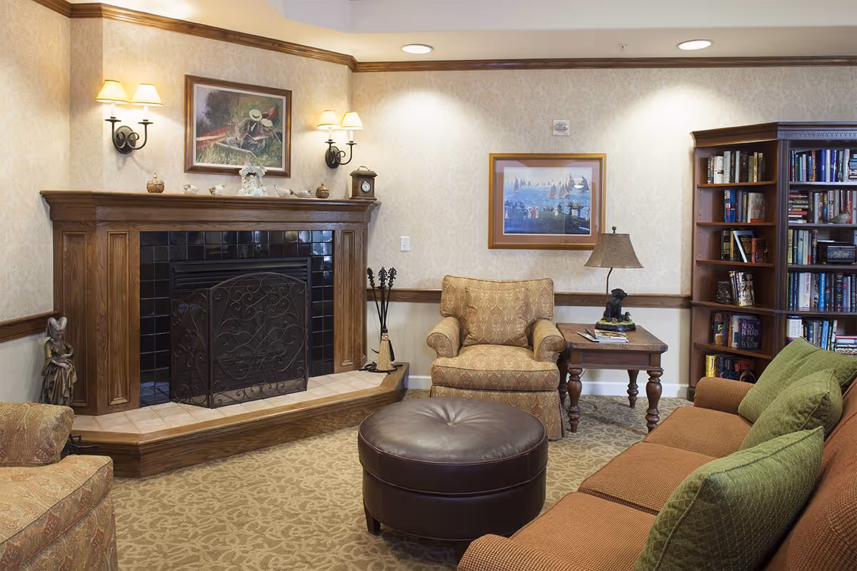 Cozy lounge with a wood-paneled fireplace, upholstered chairs, a sofa with green pillows, a round leather ottoman, and a bookshelf.