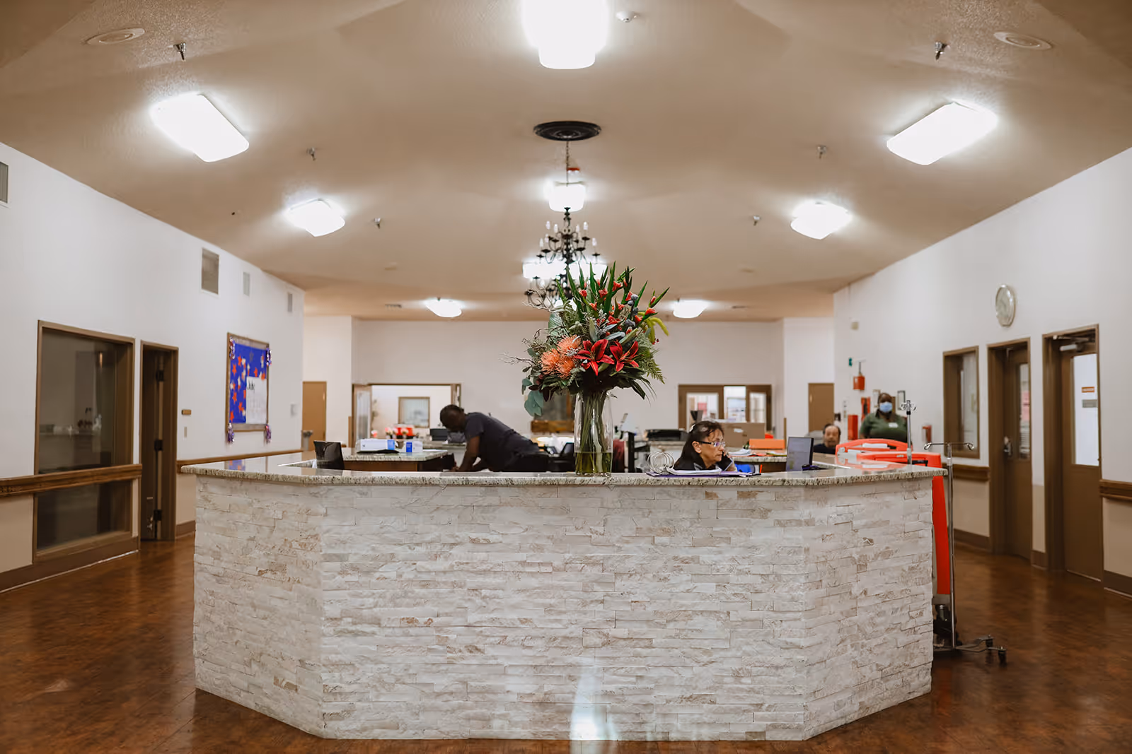 Reception area inside Cross Timbers Rehabilitation & Healthcare Center with a large stone front desk, a vase of flowers on the desk, and staff members working behind it. The room has white walls, wooden flooring, multiple doors, and ceiling lights.