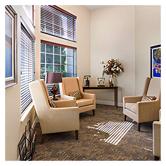 A cozy sitting area with four beige armchairs arranged around a corner. The room has large windows with blinds allowing natural light to cast shadows on the carpeted floor. A small table against the wall holds decorative items including a vase with dried branches. Framed artwork is visible on the walls.