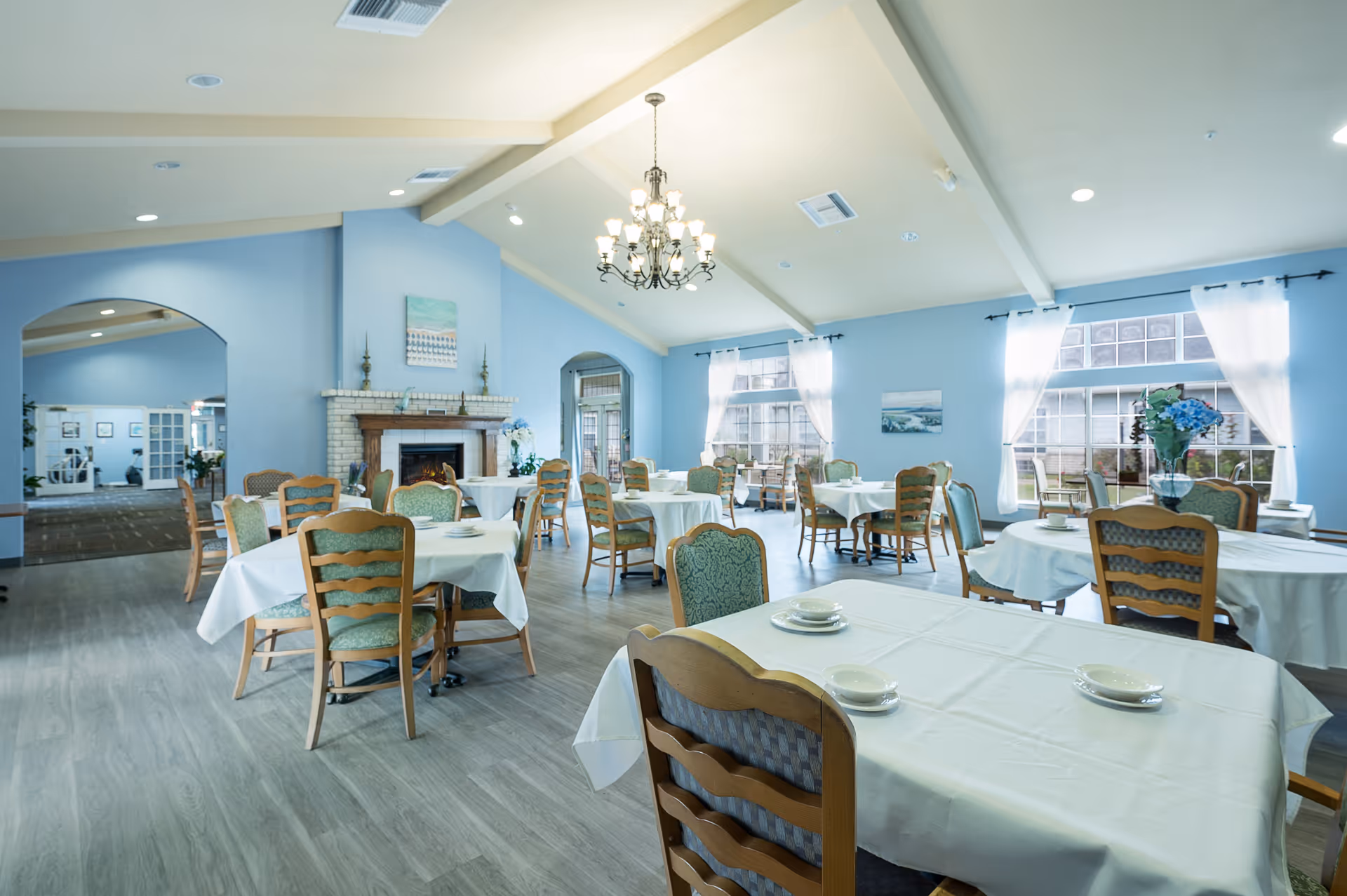 A bright dining room with multiple round tables covered with white tablecloths, each set with plates and cups. Wooden chairs with green upholstered seats surround the tables. The room features light blue walls, large windows with white curtains, a chandelier hanging from a vaulted ceiling, and a fireplace with decorative items on the mantel. An open archway leads to another room in the background.
