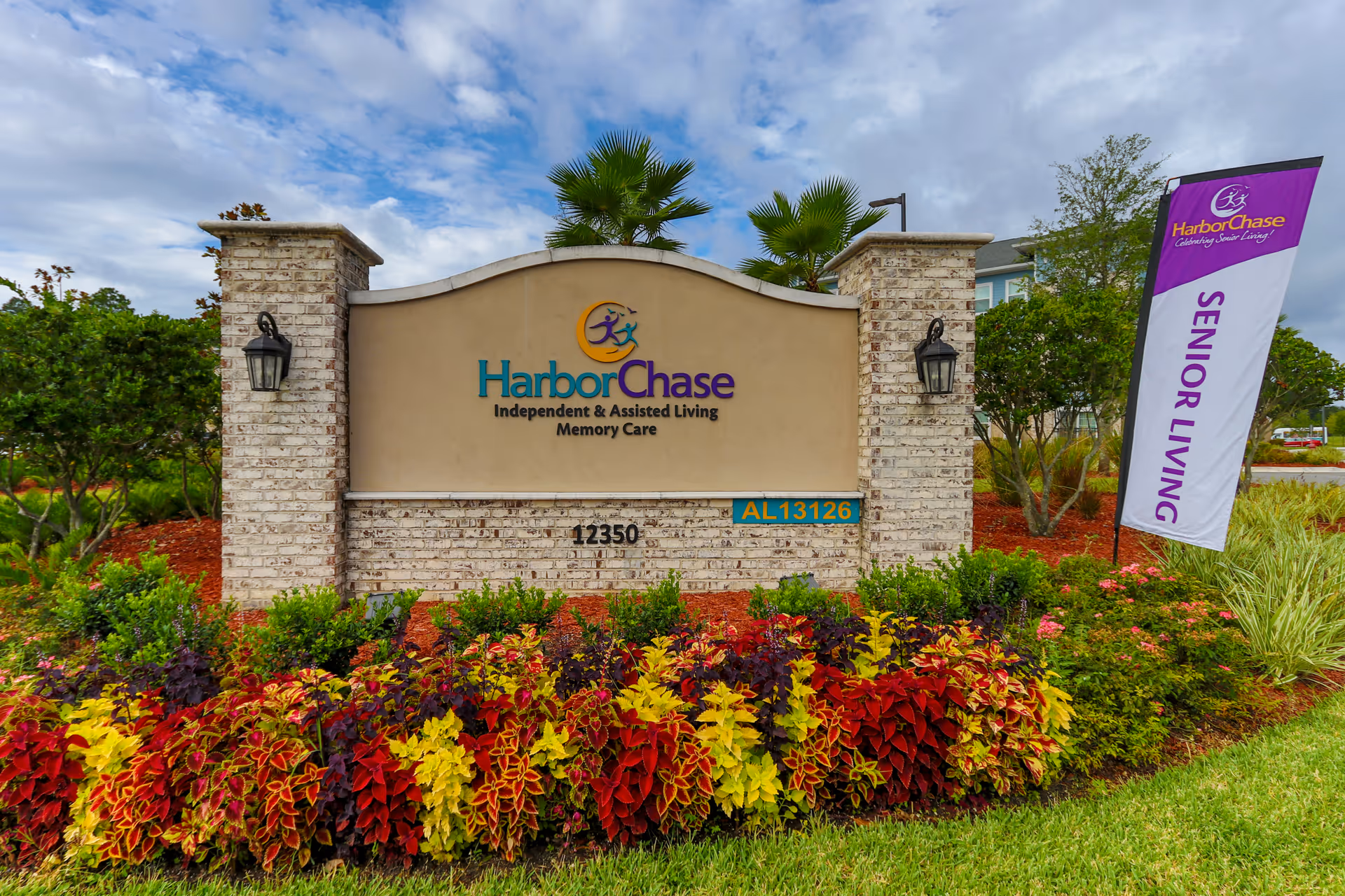 Entrance sign for HarborChase, an independent and assisted living memory care facility, surrounded by colorful landscaping with red, yellow, and green plants, two brick pillars with lanterns, and a purple and white flag that reads 'HarborChase Senior Living'.