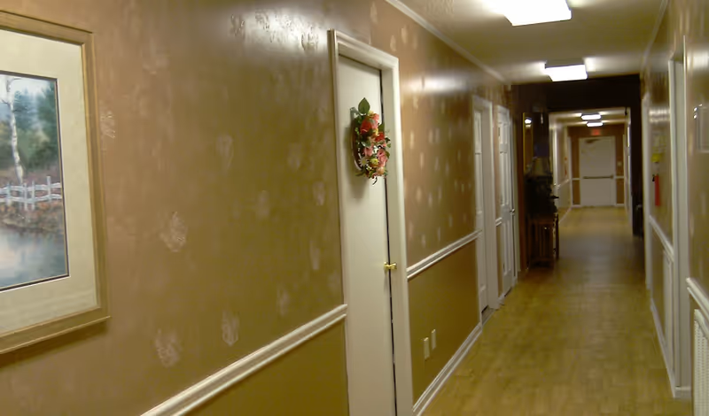 A long indoor hallway with light brown patterned wallpaper and wooden flooring. Several white doors line the hallway, one decorated with a floral wreath. A framed painting hangs on the left wall, and there is a small table with a lamp further down the corridor.