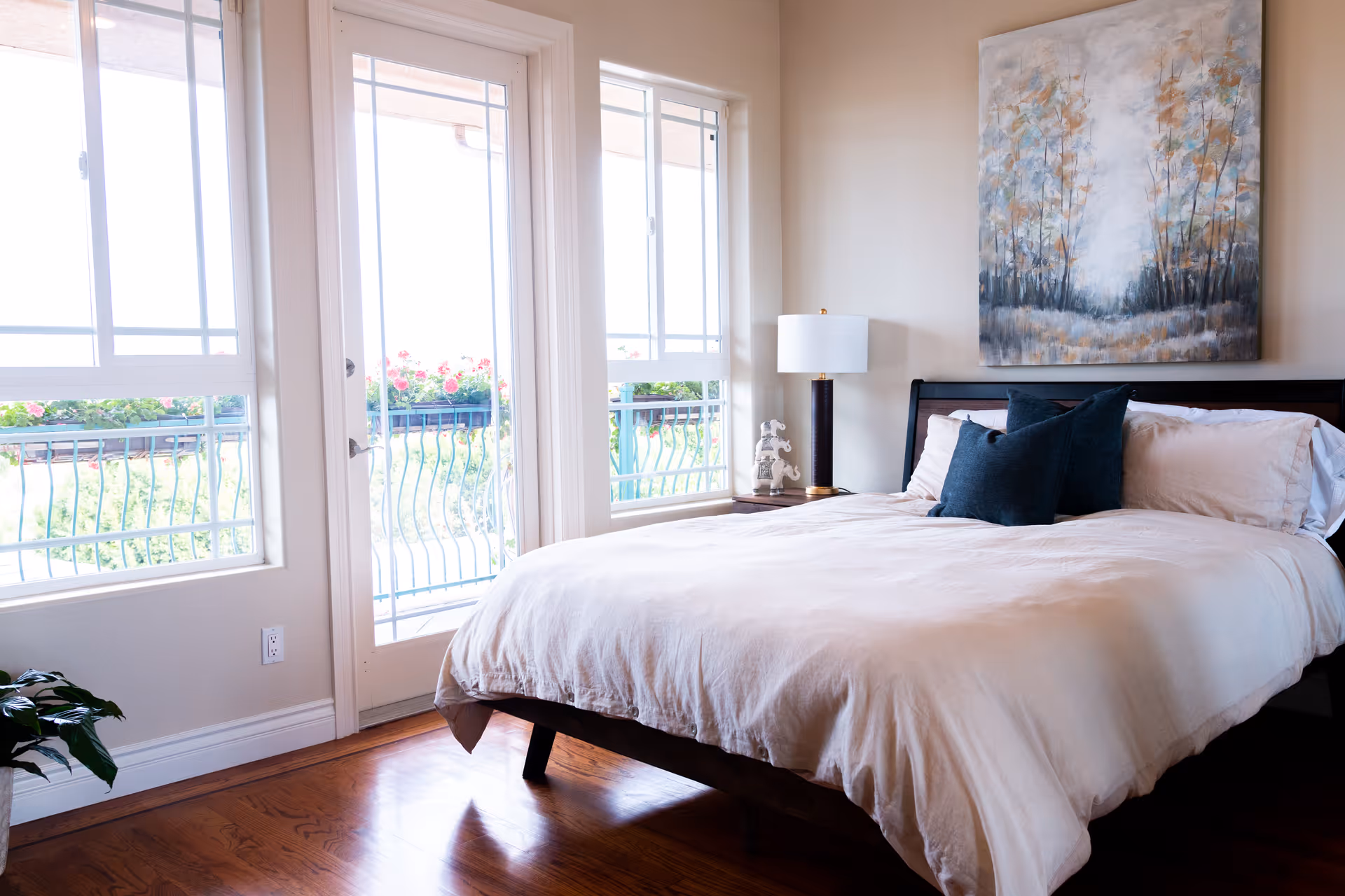 A bright bedroom with a large bed covered in white bedding and two dark blue pillows. There is a wooden nightstand with a lamp and decorative item next to the bed. Large windows and a glass door with white frames let in natural light and show a balcony with a railing and flowers outside. A painting of trees hangs on the wall above the bed.