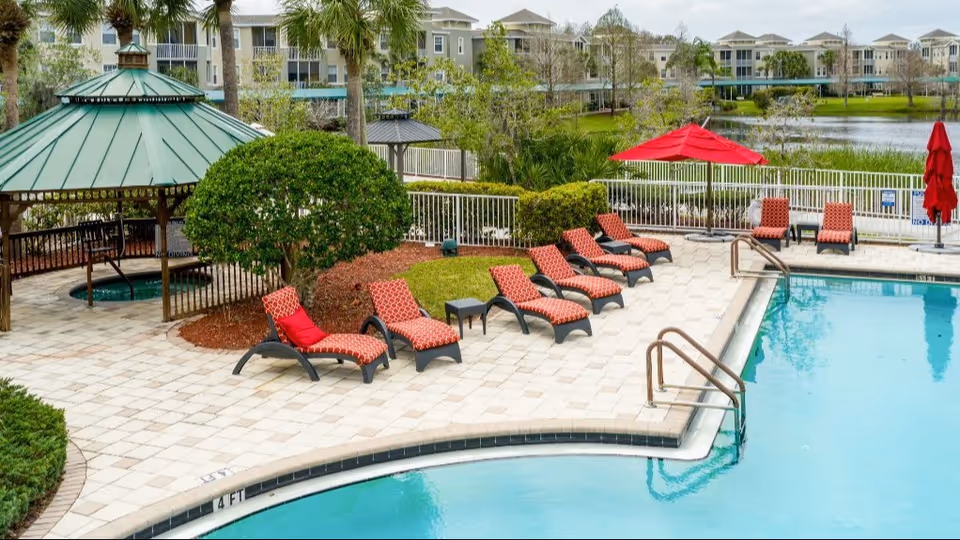 Outdoor swimming pool area with multiple red cushioned lounge chairs arranged along the poolside. A green-roofed gazebo with a hot tub underneath is visible on the left side. In the background, there are trees, a lake, and multi-story residential buildings.