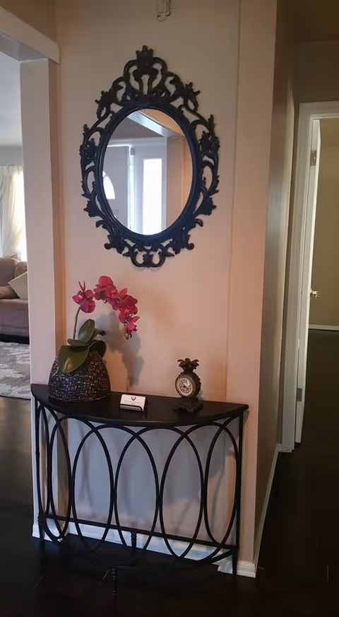 A decorative black metal console table with a woven basket holding a pink orchid and a small pineapple-shaped clock on top. Above the table is an ornate black oval mirror hanging on a beige wall. The scene is part of an interior hallway with doorways visible on the right and a glimpse of a living room area on the left.