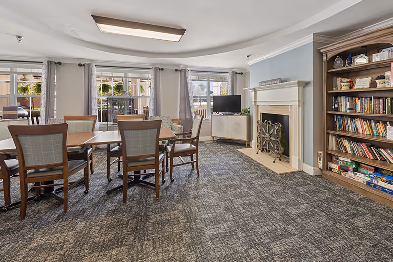A cozy common area with a carpeted floor, several wooden tables and chairs, large windows with curtains letting in natural light, a fireplace with a decorative screen, a TV on a cabinet, and a wooden bookshelf filled with books and board games.