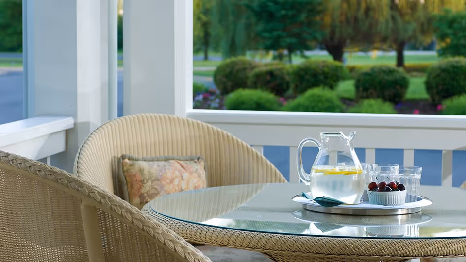 Outdoor patio area with a round glass-top wicker table and two wicker chairs, one with a floral cushion. On the table is a tray holding a glass pitcher of water with lemon slices, three empty glasses, and a small bowl of cherries. In the background, there is greenery including bushes and trees.
