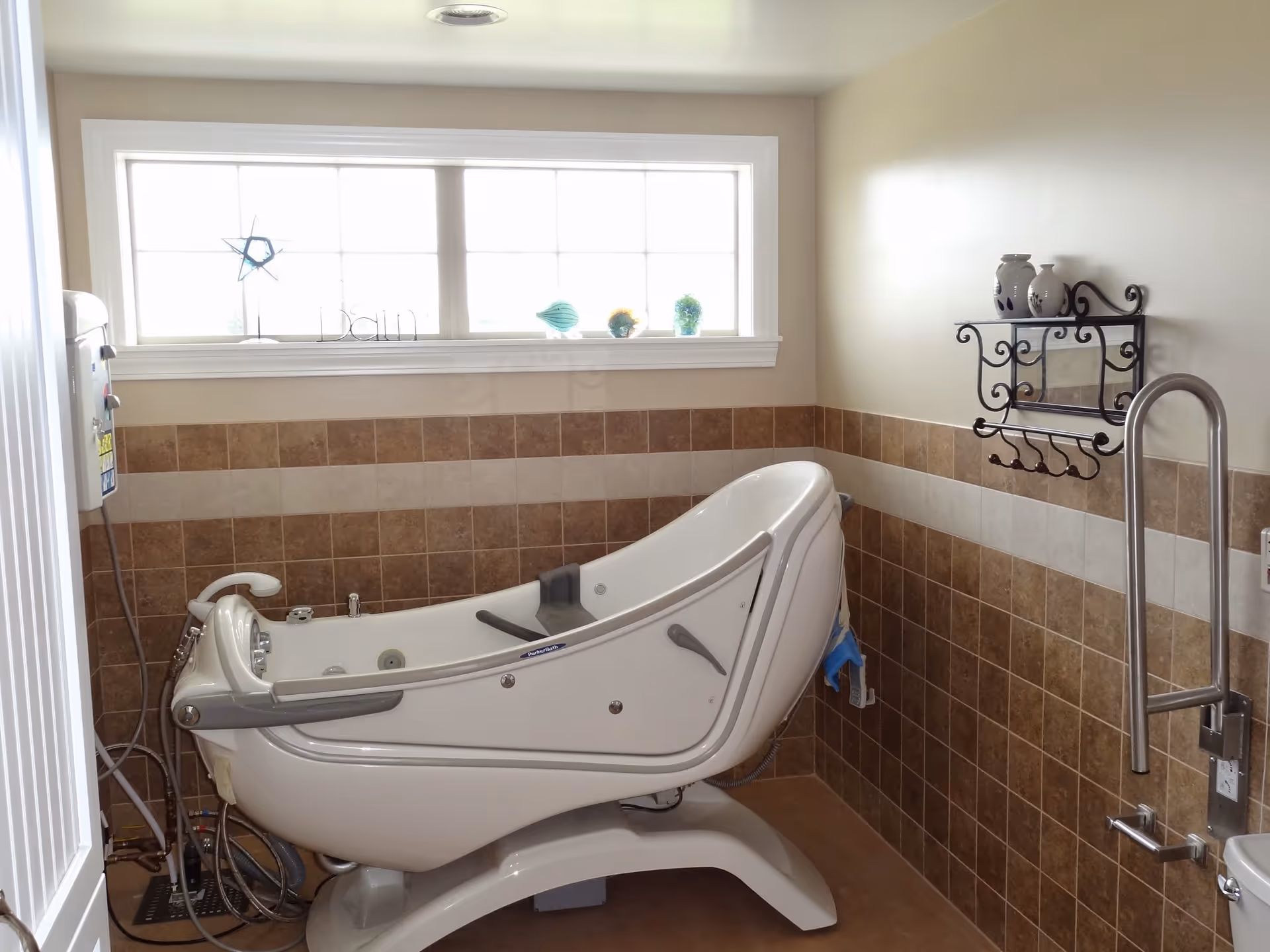 A bathroom with a large, white, medical-style bathtub designed for assisted bathing. The walls are tiled halfway up with brown and beige tiles, and there is a window above the tub letting in natural light. A decorative black metal shelf with hooks is mounted on the wall to the right, holding two ceramic jars. There is also a metal grab bar near the tub for support.