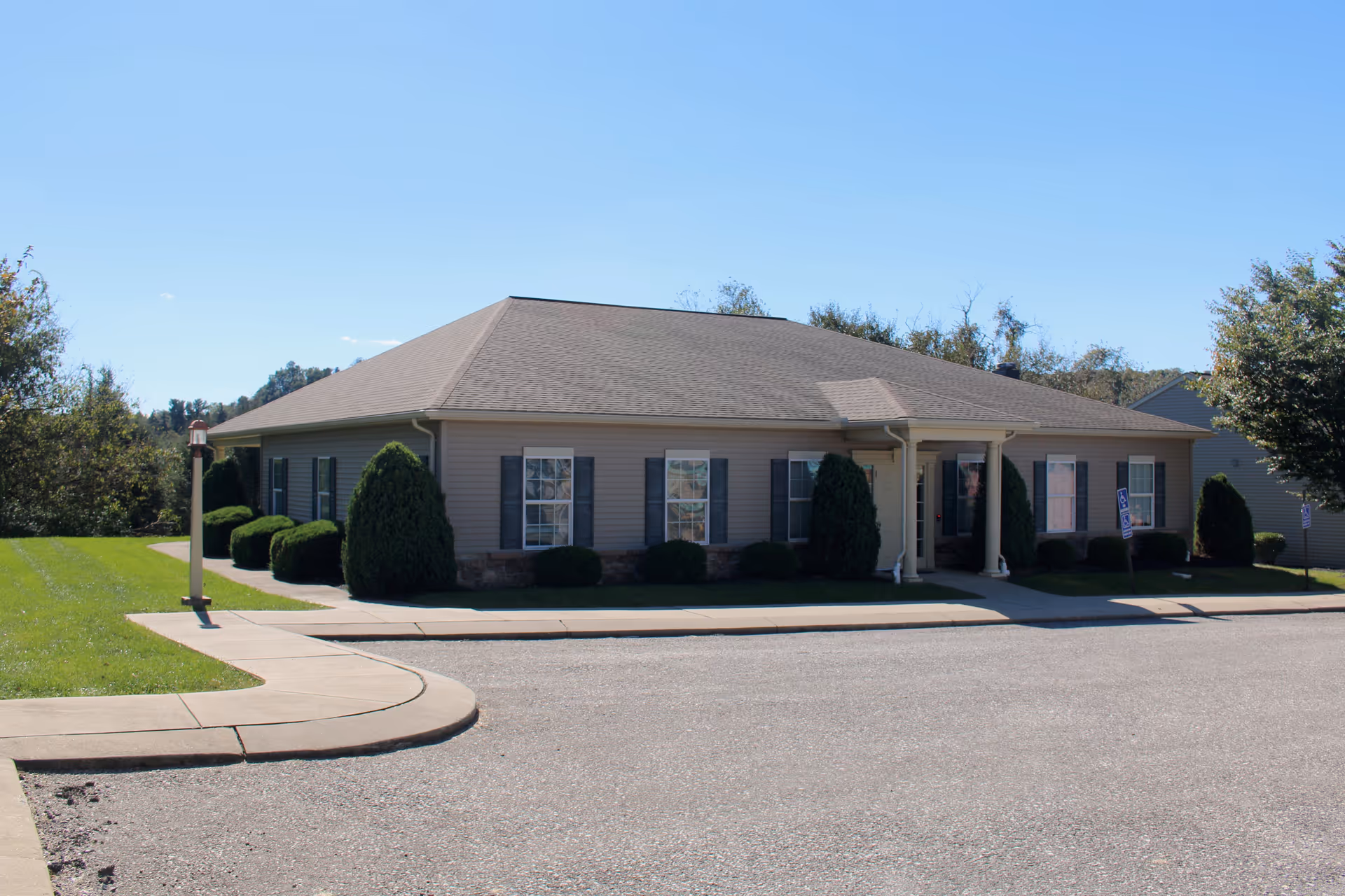 Single-story building with beige siding and a gray shingled roof, surrounded by neatly trimmed bushes and a green lawn under a clear blue sky. A sidewalk curves around the front of the building leading to a covered entrance with white columns.
