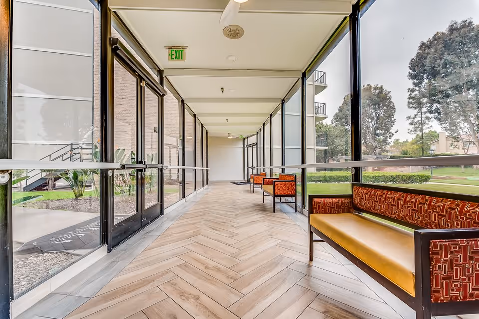 A bright indoor corridor with large floor-to-ceiling windows on both sides, featuring wooden herringbone-patterned flooring and several orange and yellow cushioned benches along the right side. Outside the windows, there is green grass and tall trees visible. An exit sign is mounted on the ceiling near the glass door on the left.