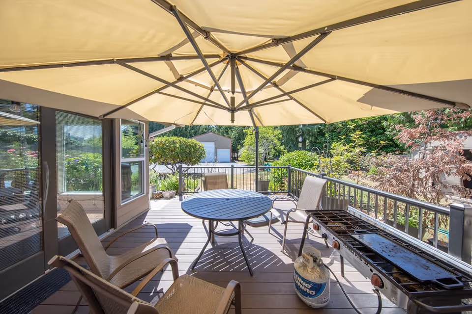 Outdoor patio area with a large beige umbrella providing shade over a round table and several chairs. There is a propane grill on the right side and greenery including trees and bushes surrounding the patio. A building and driveway are visible in the background.