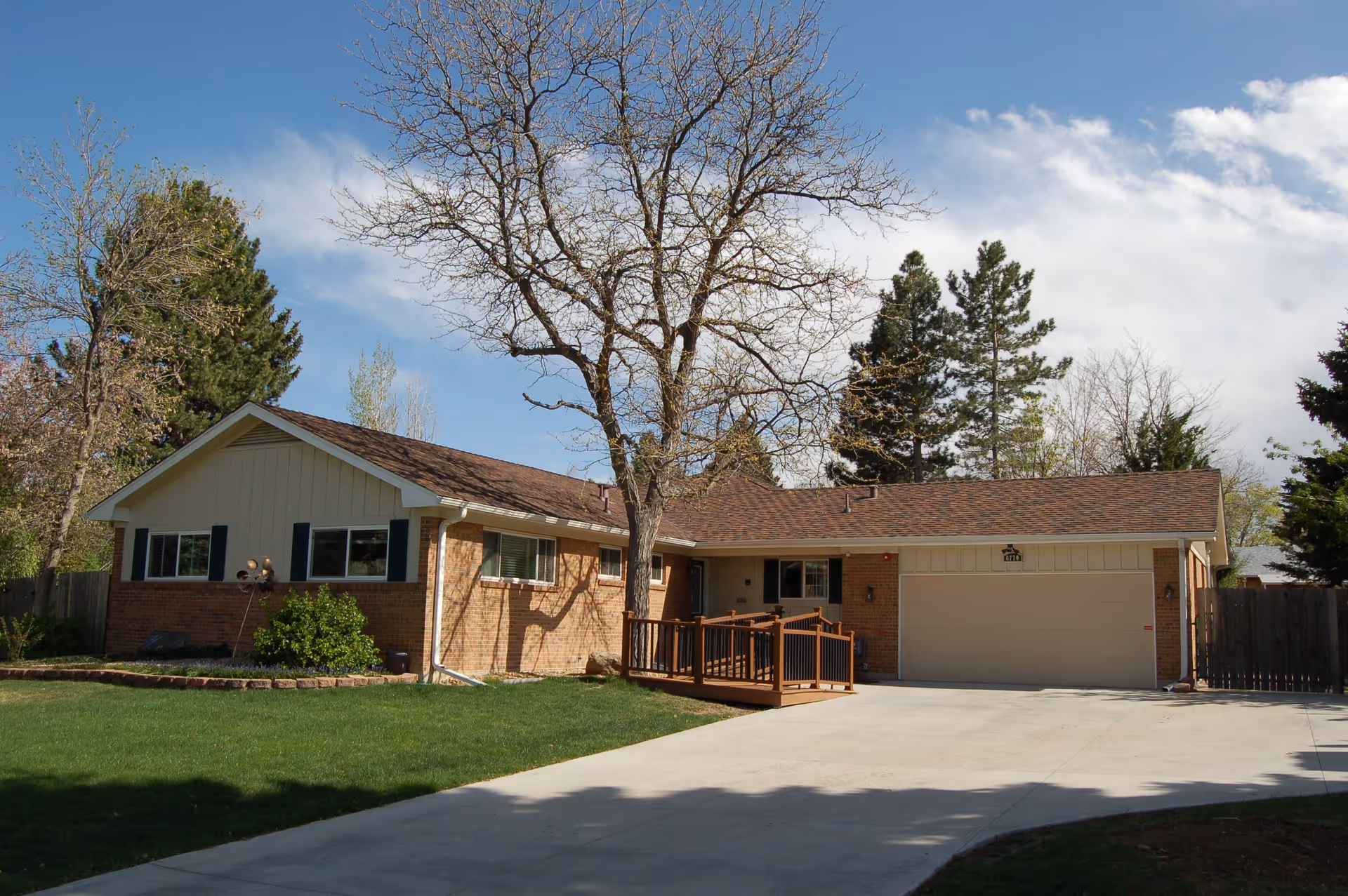 Single-story brick house with a brown shingled roof, a large tree with no leaves in front, green lawn, and a concrete driveway leading to a two-car garage. The house has a wooden ramp leading to the front door. The sky is partly cloudy with patches of blue.