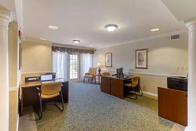 Carpeted community room with desks, chairs, desktop computers, a lamp and glass French doors letting in natural light.