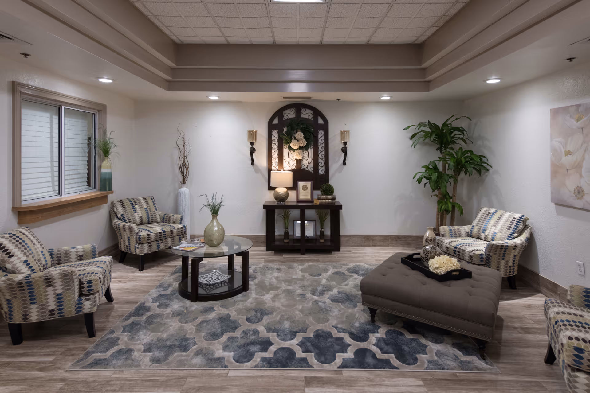 A furnished seating area with patterned armchairs, an upholstered ottoman, coffee table, rug, console table and decorative plants and wall decor.