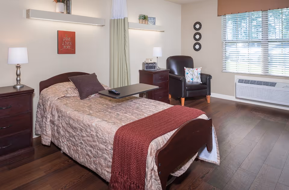 A cozy bedroom in a senior living facility featuring a single bed with a beige quilt and a red throw blanket. The bed has a dark wood headboard and footboard, with a small overbed table holding a remote control. On either side of the bed are dark wood nightstands with lamps. A black leather armchair with a floral pillow sits near a window with blinds, and the room has wood flooring. Wall decorations include a red sign that reads 'LOVE LIVES HERE' and three circular black wall hangings.