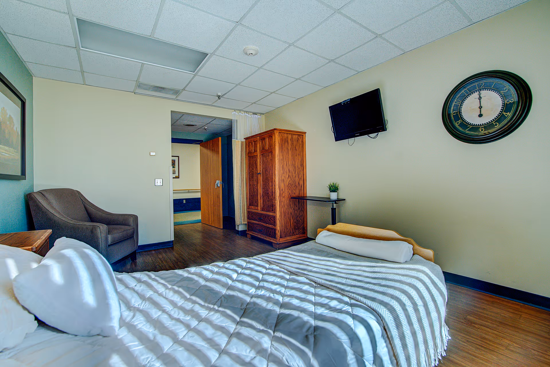 A well-lit bedroom in a senior living facility featuring a bed with white bedding and pillows, a wooden armoire, a small table with a potted plant, a wall-mounted flat screen TV, a large round clock on the wall, a gray armchair, and a framed picture. The room has wood flooring and a drop ceiling with fluorescent lighting.