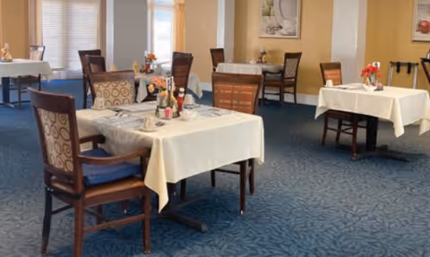 Dining room with several tables covered in white tablecloths, wooden chairs, and small floral centerpieces on a patterned blue carpet.