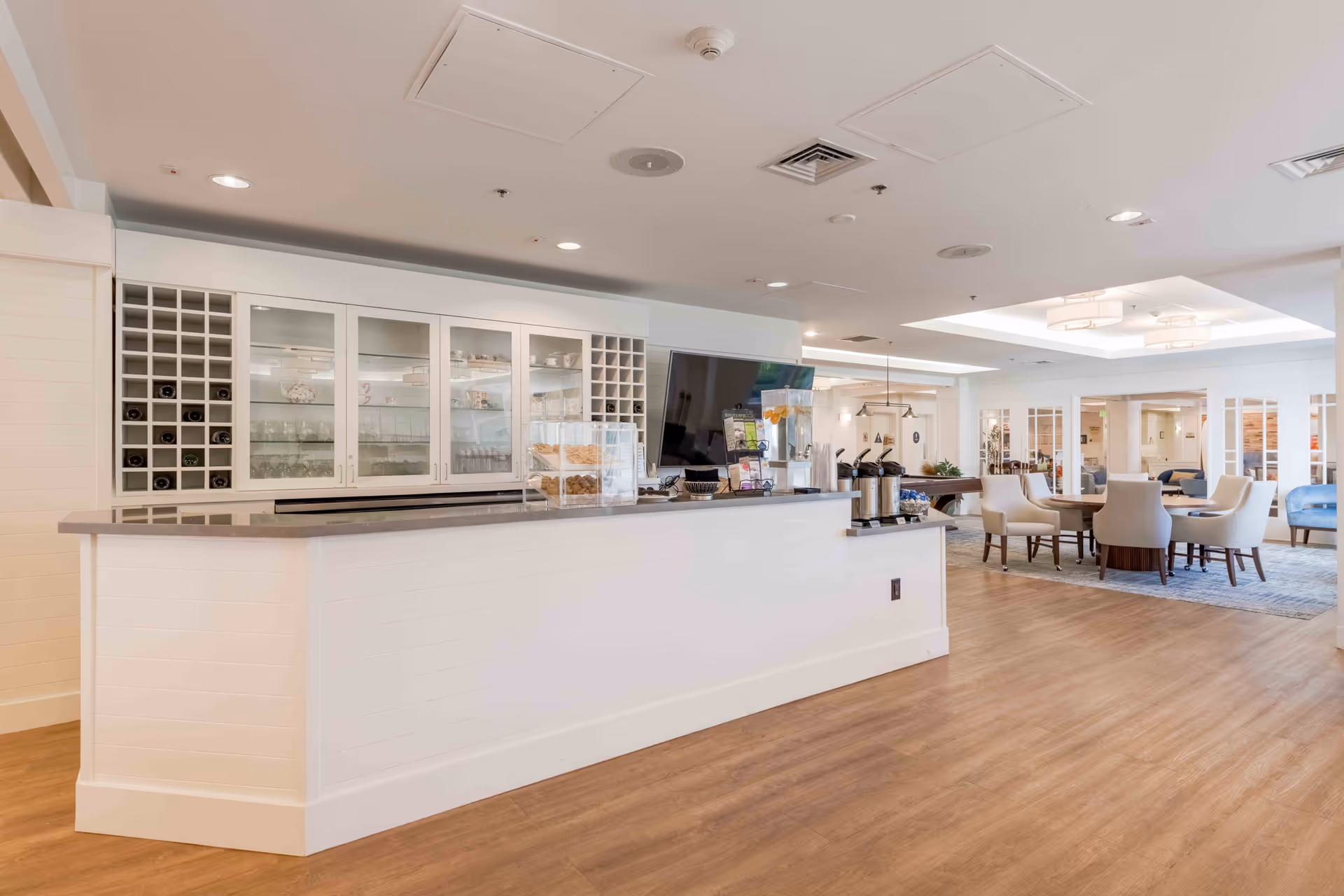 A bright and spacious common area in a senior living facility featuring a long white counter with a granite top, glass cabinets with glassware, wine racks, a large flat-screen TV, and a seating area with round tables and cushioned chairs. The room has wood flooring and recessed lighting in the ceiling.