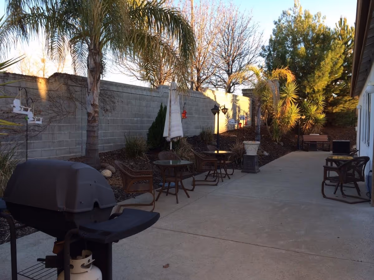 Outdoor patio area with a concrete floor, several small round tables with chairs, a barbecue grill, palm trees, and other greenery along a cinder block wall. There is a bench and some decorative lamp posts along the pathway next to a building.