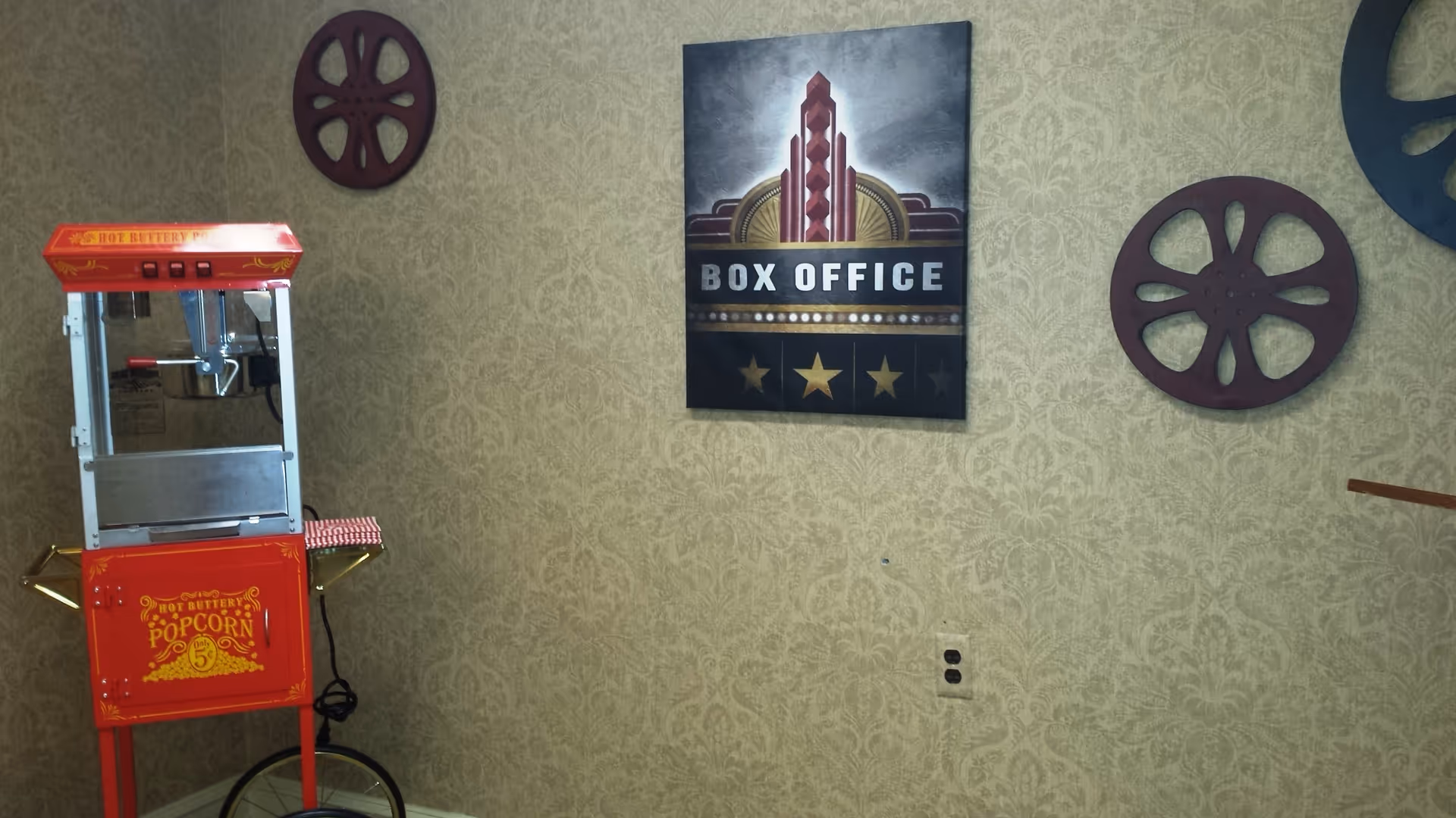 A vintage-style red popcorn machine with the text 'Hot Buttery Popcorn only 5¢' stands against a patterned beige wall. Above and to the right of the machine are decorative film reels and a framed sign that reads 'BOX OFFICE' with a stylized theater marquee design.