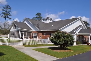Exterior view of a single-story building with white siding and brick accents, surrounded by a white picket fence and green lawn with a small tree in front. The sky is clear with a few clouds and tall pine trees are visible in the background.