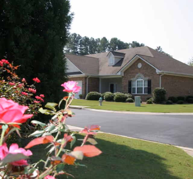 A single-story brick building with a gabled roof, surrounded by a well-maintained lawn and bushes. In the foreground, there are pink flowers and green foliage. The sky is clear and trees are visible in the background.