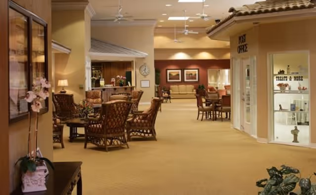 Spacious, warmly lit senior living facility interior with seating areas, tables, a small shop window labeled 'TREATS & MORE' and a 'POST OFFICE' sign.