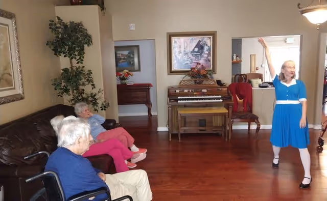 A woman in a blue dress is standing and raising one arm in a living room area with wooden floors. Two elderly people are seated on a dark brown leather couch and a wheelchair, watching her. The room has a piano against the wall, framed pictures, a plant, and a chair with a red cover.