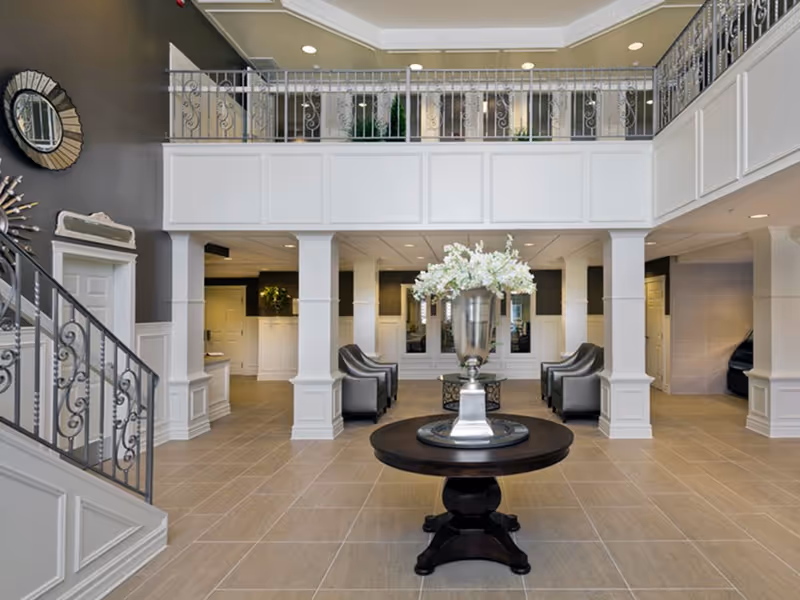 Spacious and elegant assisted living facility lobby with a round wooden table in the center holding a large silver vase filled with white flowers. The room features beige tiled flooring, white paneled columns, a decorative wrought iron railing on the upper level, and several gray armchairs arranged in seating areas. The walls are painted in neutral tones with decorative mirrors and wall art.