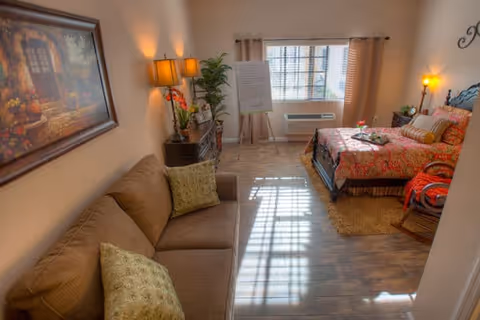 A cozy senior living room and bedroom combination featuring a brown sofa with green pillows on the left, a wooden dresser with a lamp and plants, a large window with beige curtains letting in natural light, and a bed with a colorful floral bedspread on the right side. The room has wooden flooring and warm lighting.