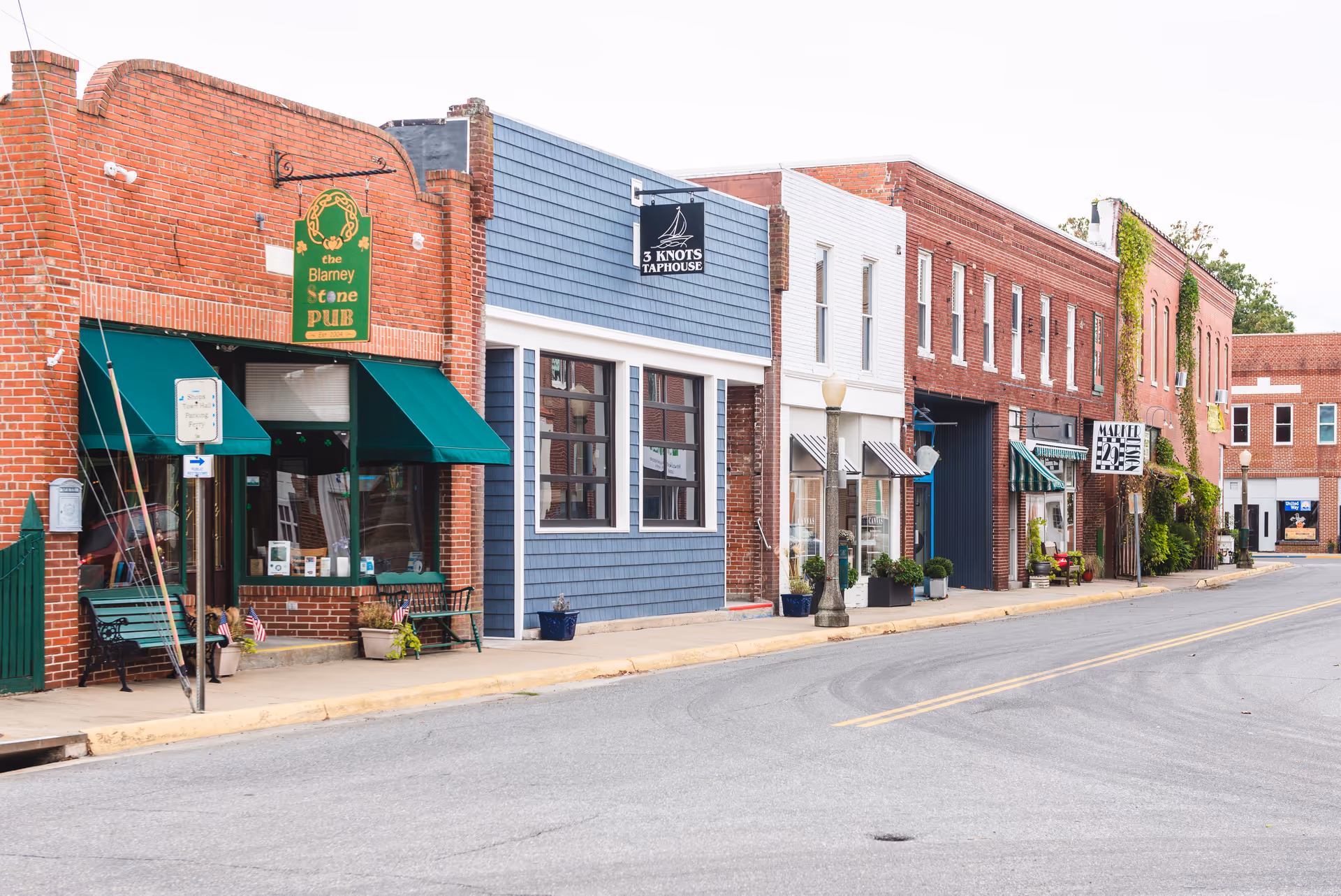 A quiet street view of a small town with a row of brick and wooden storefront buildings including the Blarney Stone Pub with green awnings and benches outside, 3 Knots Taphouse with a blue facade, and other shops with various signs and potted plants along the sidewalk.