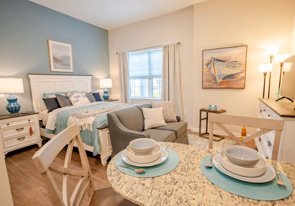 A cozy, well-lit bedroom with a white wooden bed frame, teal and gray bedding, two bedside tables with blue lamps, a small gray loveseat with pillows, a round dining table set for two with bowls and plates on teal placemats, a window with curtains, a dresser with a lamp, and a painting of a boat on the wall.