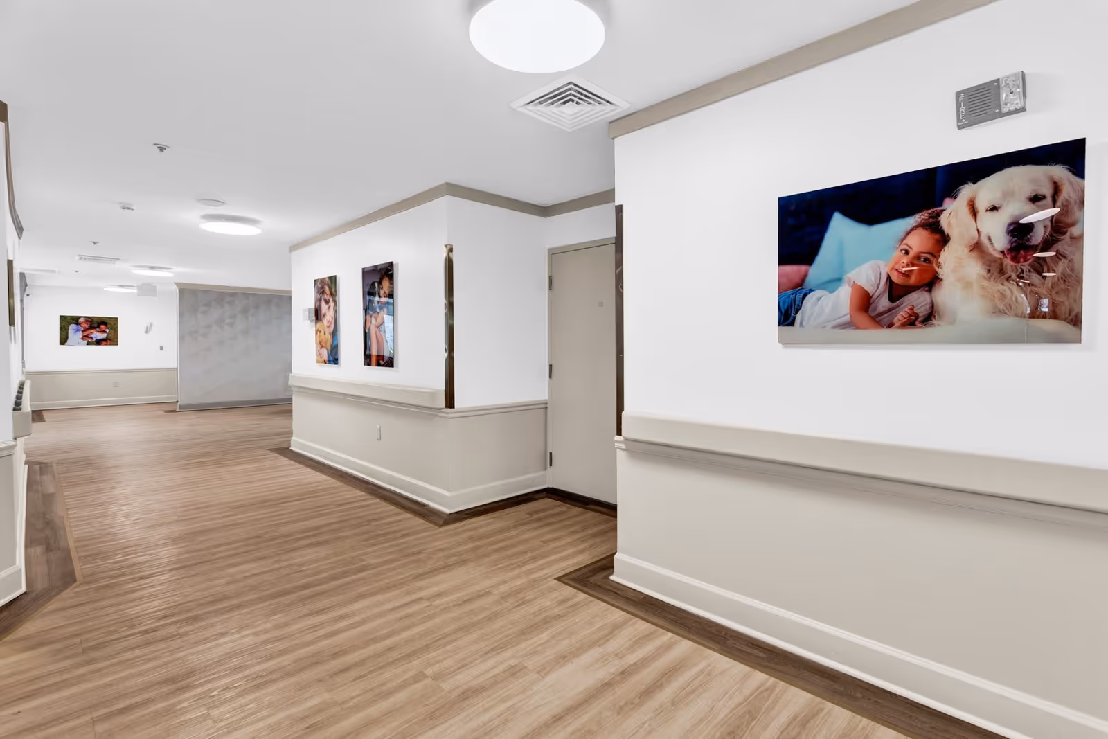 A clean, well-lit hallway in an assisted living facility with light wood flooring and white walls with beige wainscoting. The hallway features several framed photos on the walls, including images of a child with a dog and other family moments. Ceiling lights provide bright illumination.