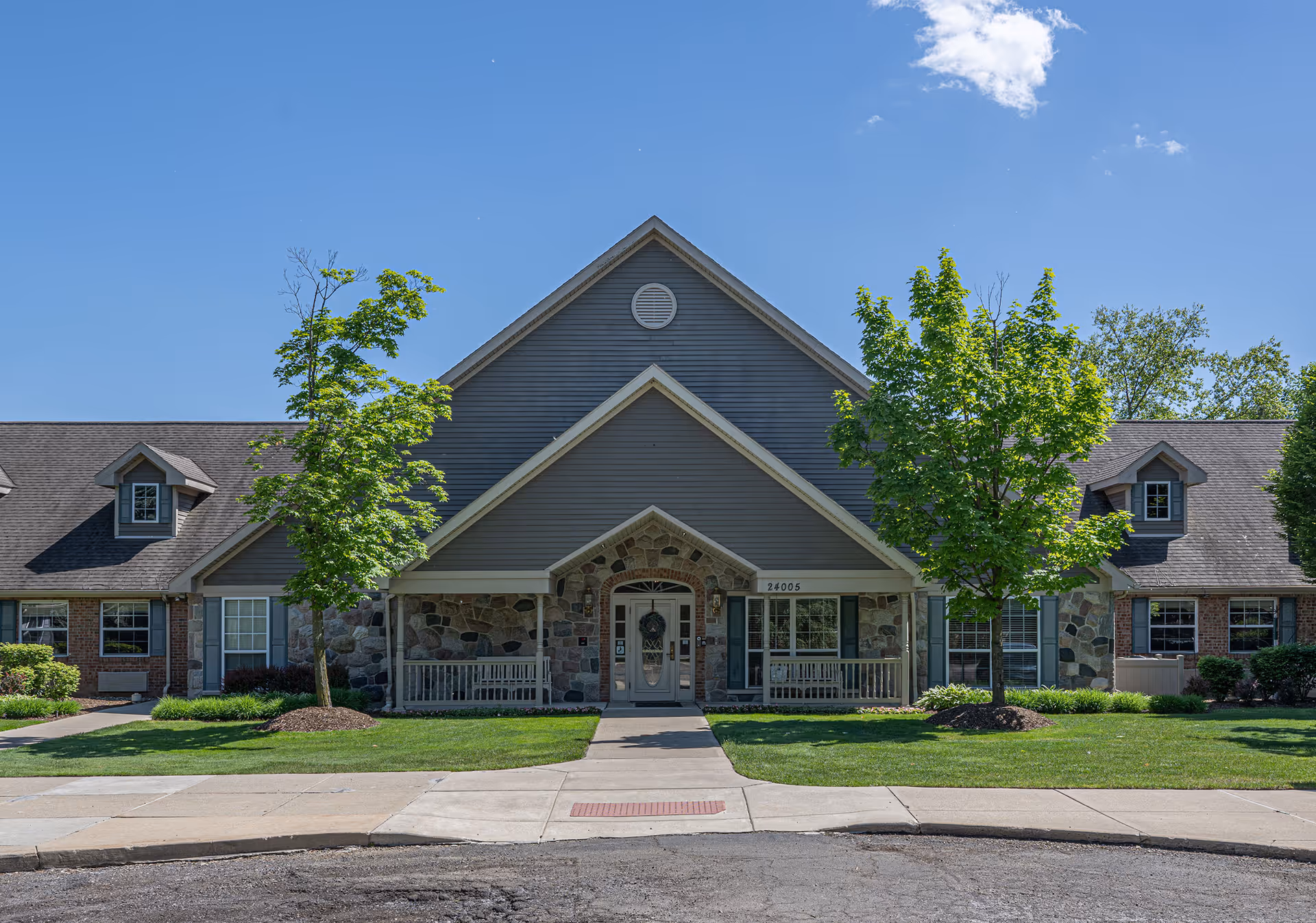 Front exterior view of Arden Courts - ProMedica Memory Care Community building with a peaked roof, stone and siding facade, two trees in front, and a sidewalk leading to the entrance door.