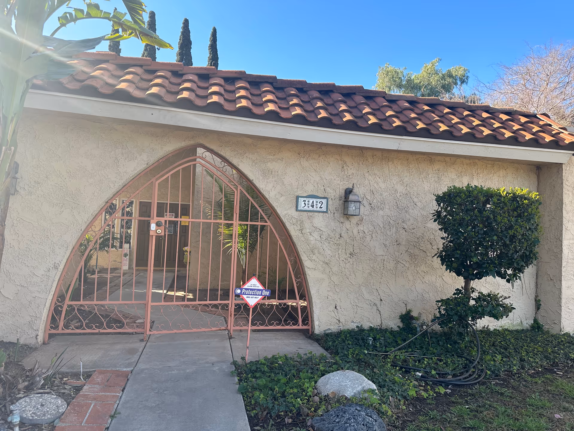 Front entrance of a stucco building with a red tiled roof and a pink arched metal gate, with address number 3442 visible.
