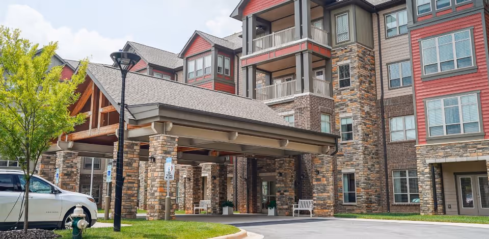 Exterior view of a senior living facility named Harmony at Greensboro featuring a covered entrance with stone pillars, a multi-story building with a mix of brick and siding, several windows, a parked white SUV, a street lamp, and a small tree on a grassy area.
